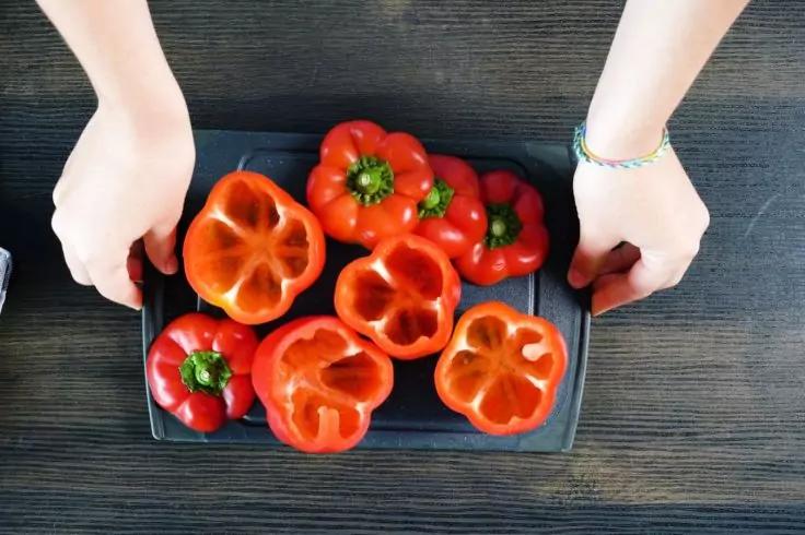 Zwei Hände halten ein schwarzes Tablett mit sechs roten Paprikaschoten, einige halbiert, auf dunklem Holzuntergrund