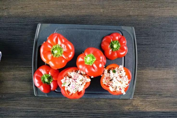 Fünf rote Paprika auf einem dunklen, rechteckigen Teller auf einem Holztisch; drei ganz, zwei halbiert und mit heller, fein zerkleinerter Füllung