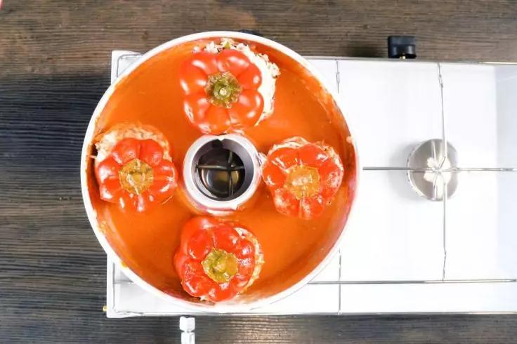 Drei rote Paprika mit der Schnittfläche nach oben in einer runden orangefarbenen Form mit Mittelrohr, rote Sauce in der Form, weiße Herdplatte rechts