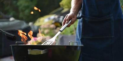 Oberk&ouml;rper einer Person mit Sch&uuml;rze, die eine Metallschaufel &uuml;ber einem Holzkohlegrill mit sichtbaren Flammen h&auml;lt