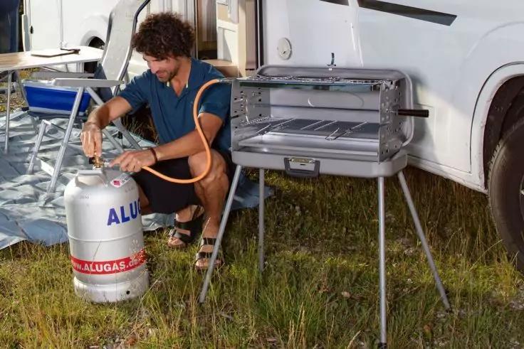 Mann kniet neben einer Gasflasche und verbindet einen Schlauch mit einem tragbaren Grill, im Hintergrund Wohnmobil und Campingst&uuml;hle auf Gras