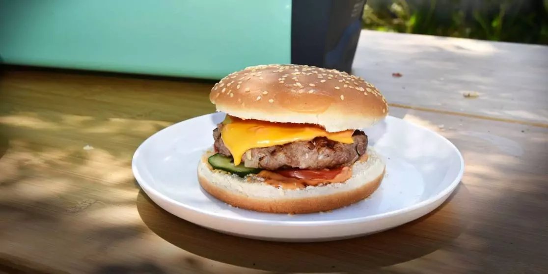 Hamburger mit Sesambr&ouml;tchen, geschmolzenem K&auml;se, Fleischpatty, Tomatenscheibe und gr&uuml;nen Gem&uuml;sescheiben auf einem wei&szlig;en Teller auf einem Holztisch im Freien