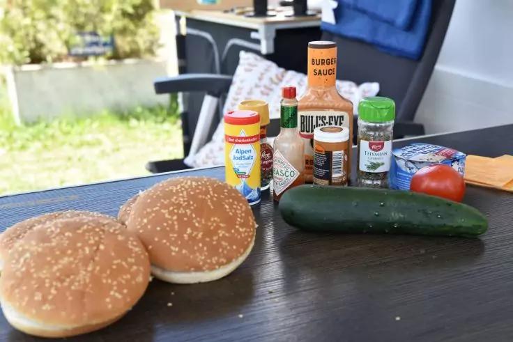 Zwei Sesambr&ouml;tchen, eine Gurke und eine Tomate sowie mehrere Gew&uuml;rz- und Saucenflaschen auf einem dunklen Tisch; im Hintergrund Garten und Stuhl