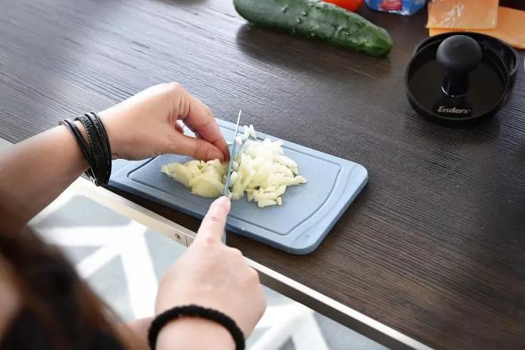 Zwei H&auml;nde schneiden eine Zwiebel auf einem kleinen Schneidebrett auf einer dunklen K&uuml;chenarbeitsplatte; im Hintergrund Gurke, K&auml;sescheiben und ein schwarzes K&uuml;chenger&auml;t