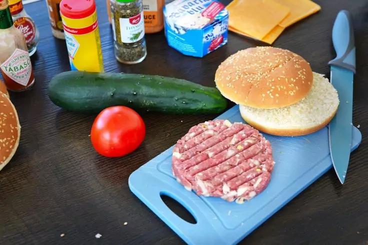 Sesambr&ouml;tchenh&auml;lften, rohes Burgerpatty auf blauem Schneidebrett, Gurke, Tomate, Messer, W&uuml;rzflaschen und verpackter K&auml;se auf dem Tisch