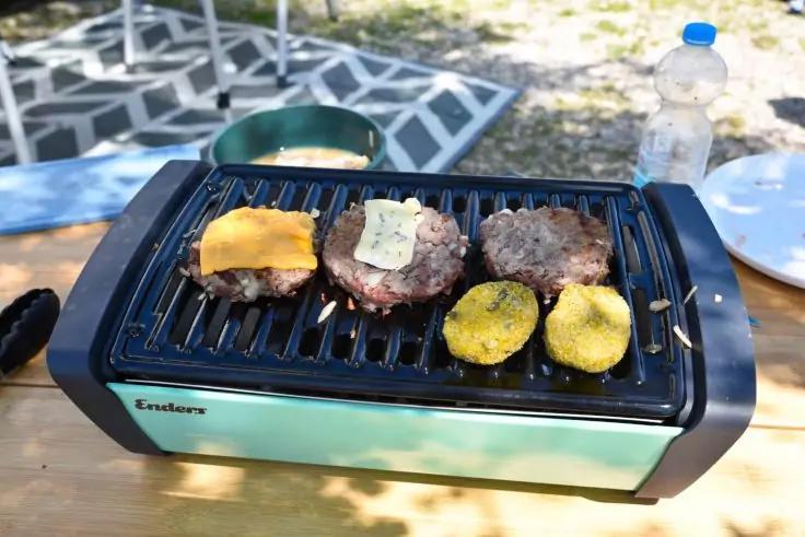 Tischgrill mit drei Burger‑Patties (eins mit geschmolzenem K&auml;se, eins mit hellem rechteckigen St&uuml;ck), zwei gelbe Gem&uuml;seh&auml;lften auf dem Rost; rechts Wasserflasche und wei&szlig;er Teller; Picknickdecke auf dem Gras im Hintergrund