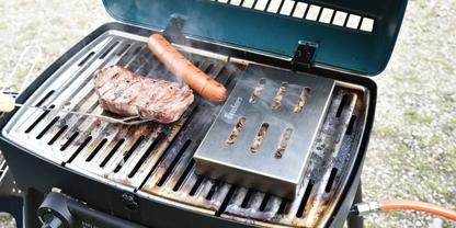 Offener Gasgrill mit Steak, Bratwurst und metallener Box mit Schlitzen auf dem Rost; Kies im Hintergrund