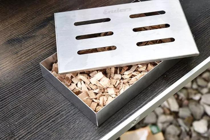 Rechteckige Metallbox mit geschlitztem Deckel, teilweise geöffnet, gefüllt mit Holzspänen, auf einer dunklen Holzfläche, daneben Kieselsteine