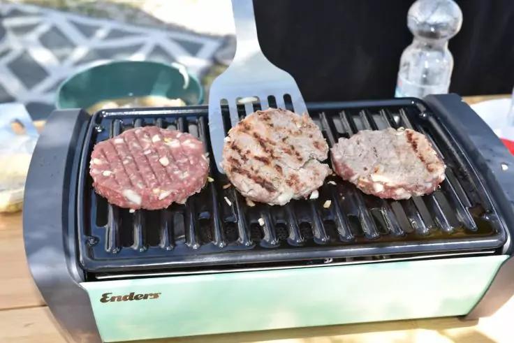 Drei Fleischpatties auf einem elektrischen Tischgrill, zwei mit Grillstreifen