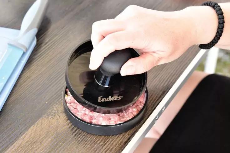 Hand mit Armband hält schwarzen Hamburgerformer mit Schriftzug "Enders" über rohem Hackfleisch in runder Form auf dunklem Holz