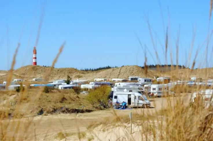 Mehrere Wohnmobile auf sandiger Fläche vor Dünen, rot-weiß gestreifter Leuchtturm im Hintergrund, Strandgras im Vordergrund