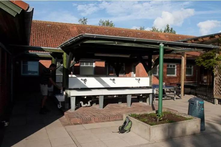 Überdachte Außenwaschstelle mit mehreren Waschbecken vor rotem Backsteingebäude, gepflasterter Hof, grüner Stützpfosten, Pflanzkübel, Rucksack auf dem Boden, eine Person links