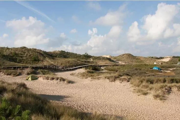 Sanddünen mit vereinzeltem Dünengras, hölzerner Bohlenweg über die Dünen, kleiner blauer Gegenstand rechts, teils bewölkter Himmel