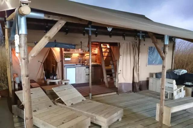 Holzterrasse mit zwei Liegestühlen unter einem überdachten Holzgestell; im Hintergrund offener Innenraum mit Küchenzeile, Vorhang und Regalen