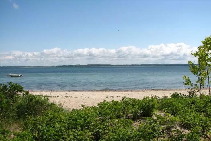 Sandstrand mit grünem Bewuchs im Vordergrund, ruhiges Meer, kleines Boot links, entfernte Küstenlinie und teilweise wolkiger Himmel