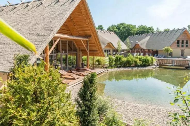 Holzhäuser mit spitzem Reetdach am Ufer eines kleinen Sees, Holzdeck mit Liegen, Büsche und Bäume im Vordergrund