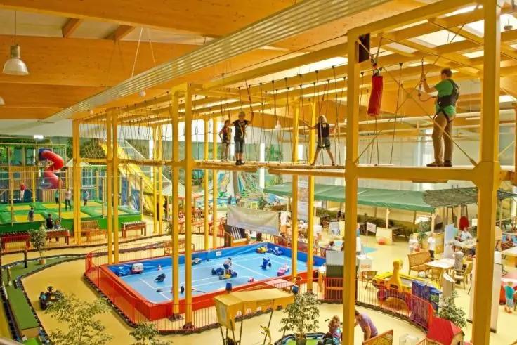 Große Halle mit gelber mehrstufiger Kletter‑ und Hochseilstruktur, Personen mit Klettergurten auf dem Seilparcours, darunter bunte Spielbereiche mit blau‑rotem aufblasbaren Becken, Rutschen und Spielgeräten