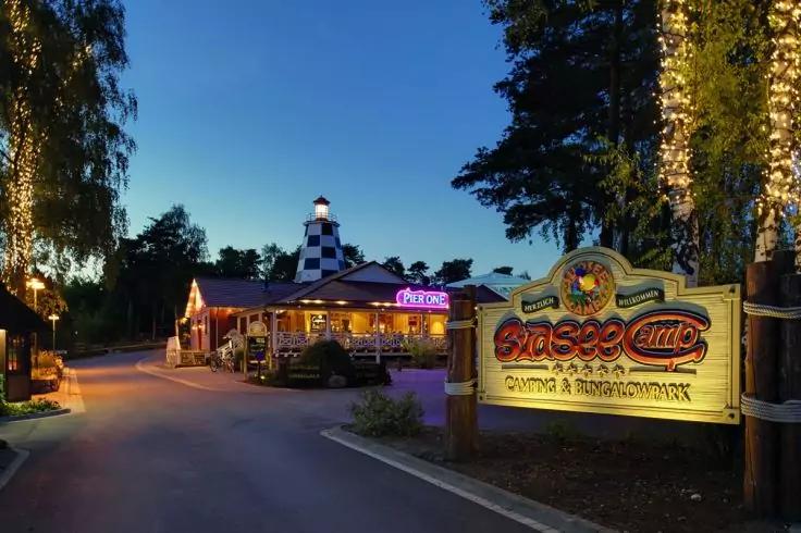 Straße bei Dämmerung, die zu beleuchteten Gebäuden und einem schwarz-weiß karierten Turm führt; rechts ein großes beleuchtetes Schild; Bäume mit Lichterketten