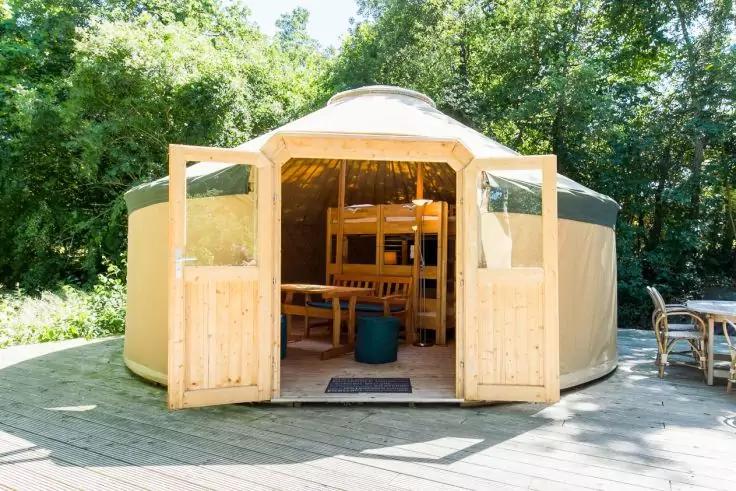 Runde Jurte mit geöffneten Doppeltüren auf einem Holzdeck; im Inneren Holztisch, Stühle, kleiner Hocker und Etagenbett; umgeben von Bäumen; rechts eine Außentischgruppe mit Stühlen