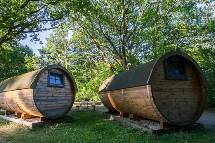 Zwei hölzerne, fassförmige Hütten mit runden Fenstern auf Stützen auf einer Wiese unter Bäumen