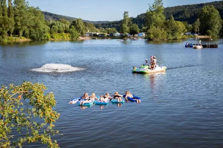 Mehrere Personen auf verbundenen Schwimmreifen auf einem See; rechts ein Tretboot mit einer Person, weiter hinten ein weiteres Boot; Ufer mit Bäumen und bewaldeten Hügeln; links eine Wasserfontäne im See