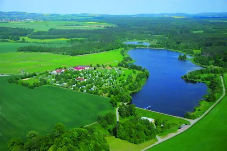 Luftaufnahme eines blauen Sees, umgeben von Bäumen, grünen Feldern, einer kleinen Gruppe von Häusern und einer Straße entlang des Ufers