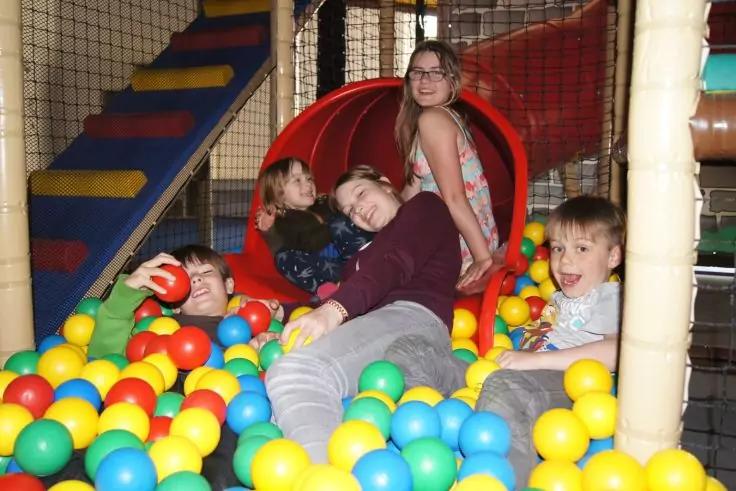 Fünf Kinder sitzen und liegen in einem bunten Bällebad vor einer roten Röhre auf einem Indoor-Spielplatz