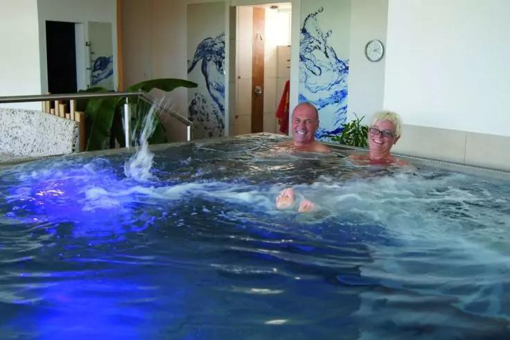 Zwei Personen in einem sprudelnden Innenwhirlpool mit blauer Beleuchtung, Wasserfontäne, Wellenmuster an der Wand, Pflanze links, offene Türen und Wanduhr