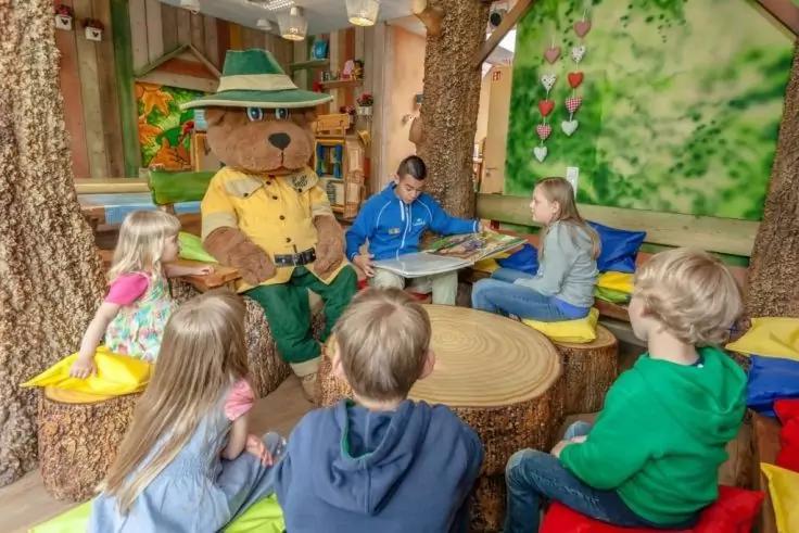 Mehrere Kinder sitzen auf bunten Kissen um einen runden Holztisch, neben einer Person im Bärenkostüm mit Hut und gelbem Hemd, Raum mit Baumstamm-Säulen und grüner Wand mit Herzdekoration