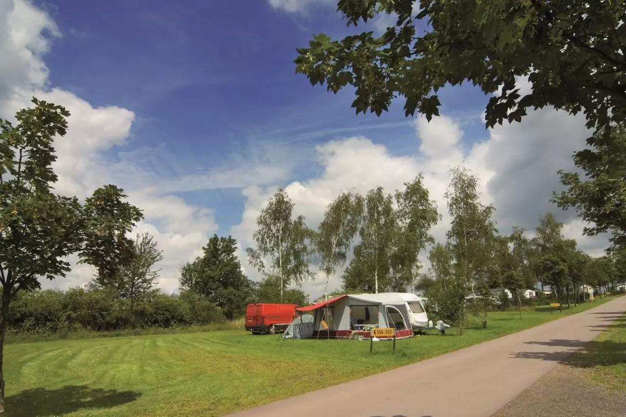 Wohnwagen mit angebautem Vorzelt auf einem Campingplatz, roter Anhänger daneben, Bäume und Wiese unter blauem Himmel mit Wolken, gepflasterter Weg rechts