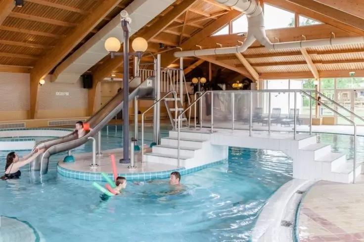 Innen-Schwimmbad mit gebogener Rutsche, Treppe und Geländern; Personen im Wasser; Holzdecke mit sichtbaren Balken; Glasabtrennung und Fenster zum Außenbereich