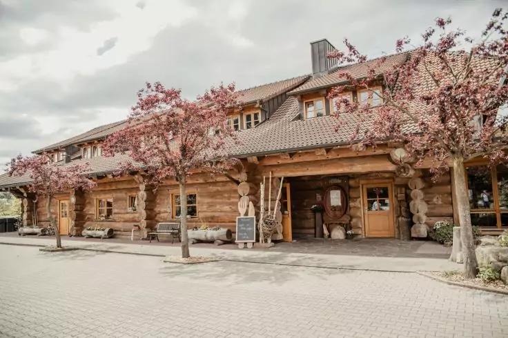 Holzgebäude mit überdachter Veranda, Fenster, Eingang und Schiefertafel; blühende Bäume davor, gepflasterter Vorplatz, bewölkter Himmel