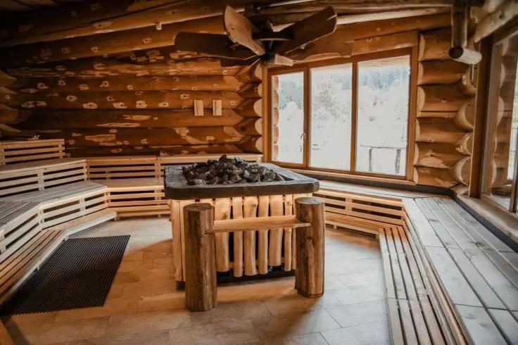 Holzsauna mit Rundholzwänden, umlaufenden Sitzbänken, zentralem Ofen mit Saunasteinen und großem Fenster