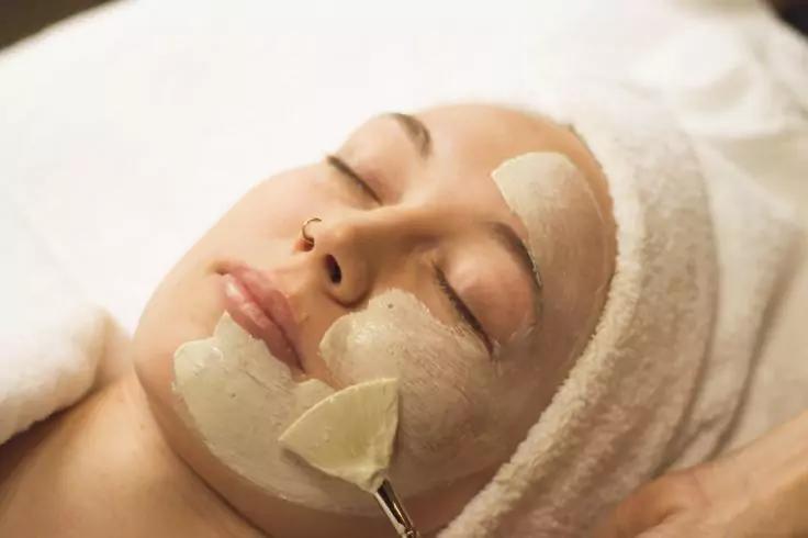 Person mit um den Kopf gewickeltem Handtuch, Augen geschlossen, Gesichtsmaske auf Wangen und Kinn, Pinsel berührt das Kinn