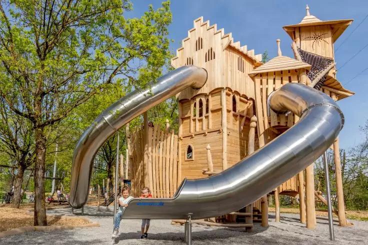 Holzspielplatz mit Turm und zwei großen gebogenen Metallrutschen, Bäume im Hintergrund, blauer Himmel, eine Person neben einer Rutsche