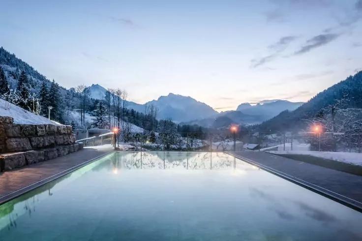 Außenpool mit ruhiger Wasseroberfläche, links eine Steinmauer, verschneite Wege und Bäume, beleuchtete Laternen am Beckenrand, schneebedeckte Berge im Hintergrund, heller Abendhimmel