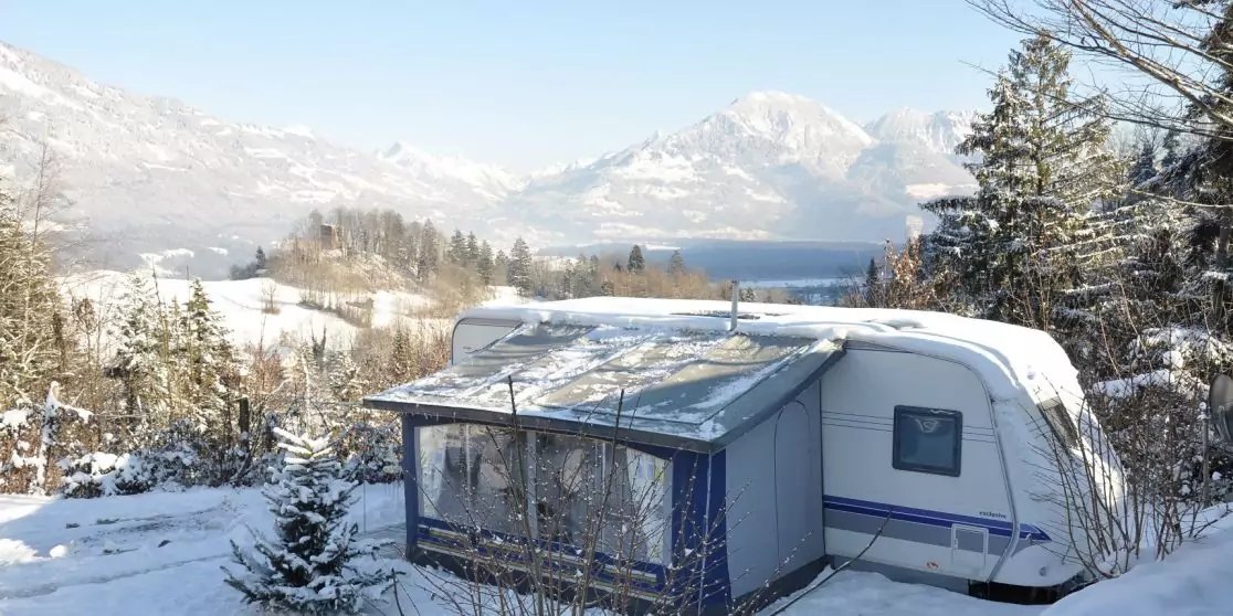 Schneebedeckter Wohnwagen mit angebautem Vorzelt auf verschneitem Boden, umgeben von B&auml;umen, mit See und Bergen im Hintergrund
