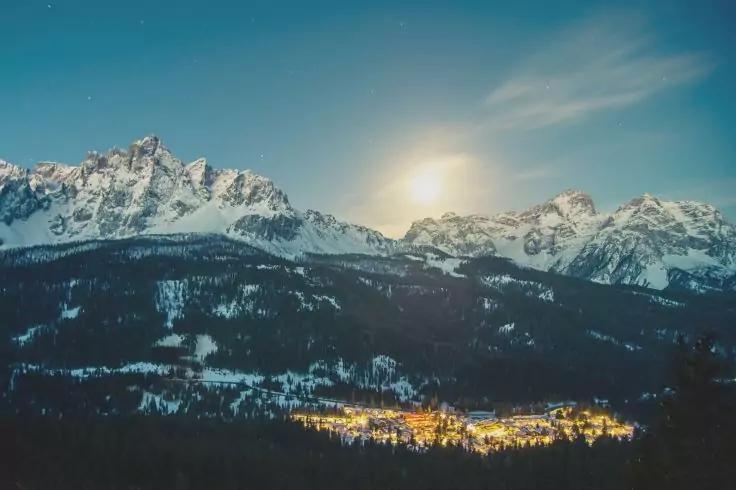 Schneebedeckte Berggipfel, leuchtender Kreis knapp &uuml;ber den Gipfeln, dunkle bewaldete H&auml;nge und ein beleuchtetes Dorf im Tal, blauer Himmel mit wenigen Wolken