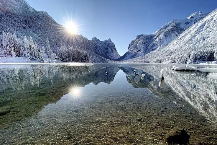 Bergsee mit klarem Wasser und sichtbarem kiesigem Grund im Vordergrund, schneebedeckte B&auml;ume und Berge, Sonne oben links, Spiegelung der Landschaft im Wasser