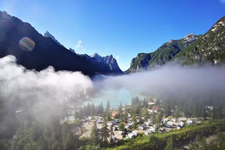 Bergsee mit Nebel &uuml;ber dem Wasser, Ufer mit B&auml;umen und kleinen Geb&auml;uden/​Fahrzeugen, steile Berge links und rechts, blauer Himmel