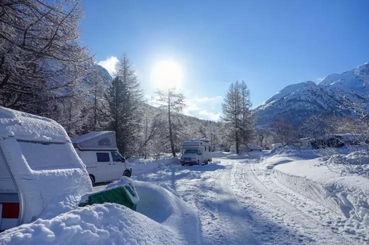 Mehrere schneebedeckte Wohnwagen auf einer verschneiten Fl&auml;che, Reifenspuren im Schnee, Nadelb&auml;ume, Berge im Hintergrund und Sonne am klaren blauen Himmel