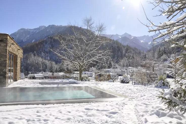 Freiluft-Schwimmbecken mit Wasser auf verschneitem Gel&auml;nde, schneebedeckte B&auml;ume, Geb&auml;ude links, Bergkette im Hintergrund und Sonne am Himmel