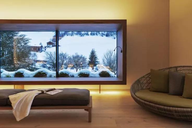 Innenraum mit gro&szlig;em rechteckigem Fenster, drau&szlig;en Schneelandschaft mit B&uuml;schen, Tannen und H&auml;usern; im Vordergrund niedrige Bank mit Decke und offenem Buch, runder Korbsessel mit Kissen, Holzboden, gelbes Licht unter dem Fenster, Stehlampe neben dem