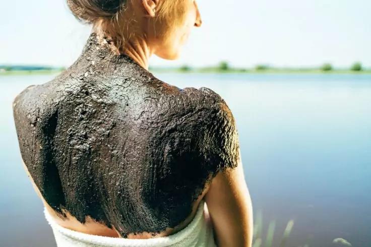 Person von hinten an einem Seeufer, Rücken und Schultern mit dunkler Schlammschicht bedeckt, Haar zusammengebunden, weißes Handtuch um die Hüften, ruhiger See im Hintergrund