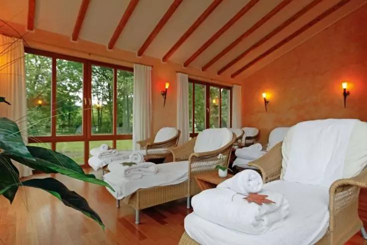 Raum mit drei geflochtenen Liegen mit weißen Handtüchern, Holzbalkendecke, großen Fenstern mit Blick auf Bäume, Wandkerzen und eine Pflanze