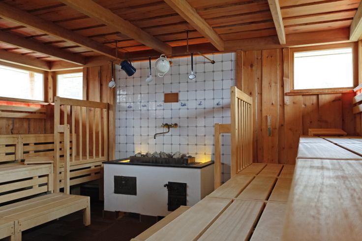 Holzverkleideter Innenraum mit gestaffelten Holzbänken, zentraler Ofen mit Steinen vor einer gefliesten Wand, schmale hohe Fenster und sichtbare Holzbalken an der Decke