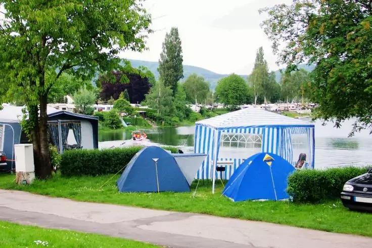 Zwei blaue Zelte und ein weiß-blau gestreifter Pavillon auf einer Wiese am See, Bäume, ein Gehweg, ein am rechten Bildrand geparkter Pkw und Hügel im Hintergrund