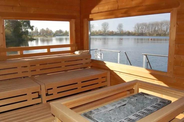 Holzinnenraum mit mehrstufigen Sitzbänken und Ofen mit Steinen; große Fenster mit Blick auf einen See und Uferbäume