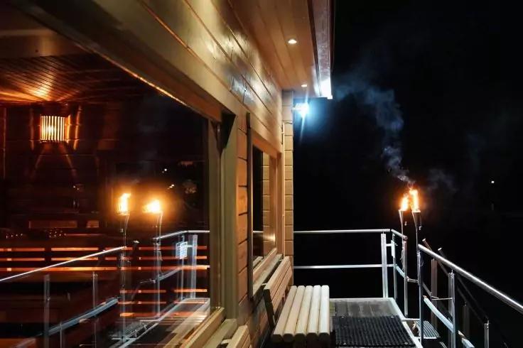 Außenwand aus Holz mit Fenster, Fensterreflexion, beleuchtete Fackeln mit Rauch an einem nächtlichen Steg, Metallgeländer und Außenlampe