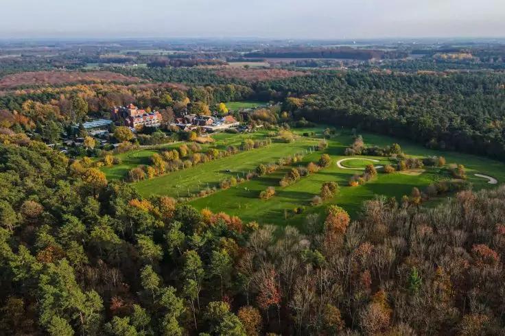Luftaufnahme eines Golfplatzes mit Fairways, Grüns, Sandbunkern, Wegen und einem Clubhaus, umgeben von Wald mit grünen, gelben und orangenen Blättern